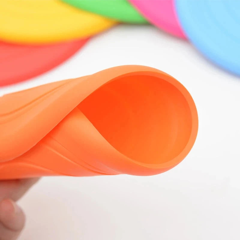 Close-up of a person holding a flexible, rolled-up, bright orange silicone disk. The background displays partially visible disks in various vibrant colors such as pink, green, yellow, and blue. The Durable Silicone Dog Flying Disc – Sturdy, Bite-Resistant Outdoor Training Toy is prominently in focus, showcasing its smooth texture and pliability.