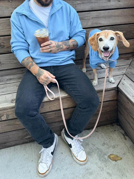 Person sitting on a wooden bench with a dog, both wearing blue sweatshirts, holding coffee and a leash.