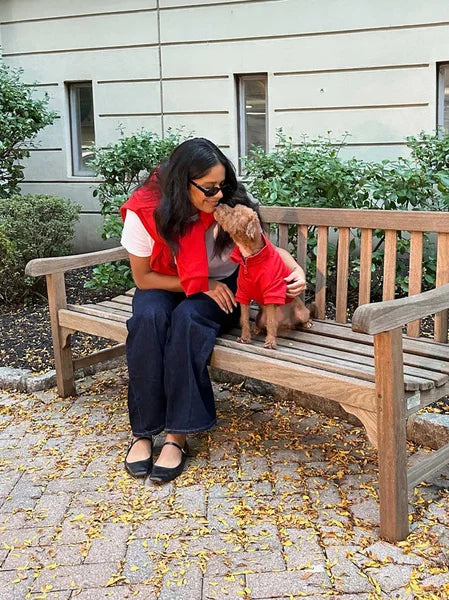 Woman sitting on a bench with a small dog, both wearing red shirts, in an outdoor setting.