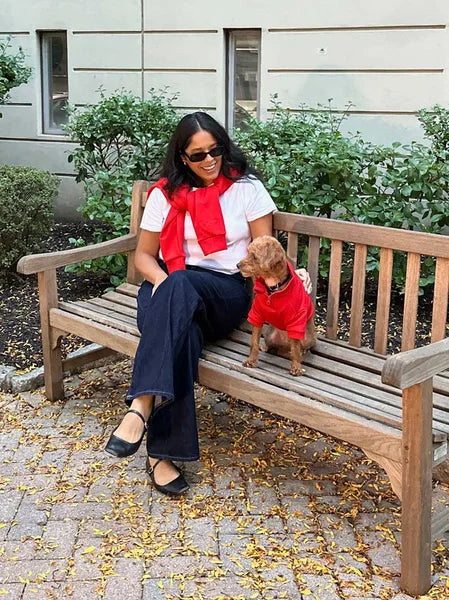 Woman sitting on a bench with a dog wearing a red sweater in an outdoor setting.