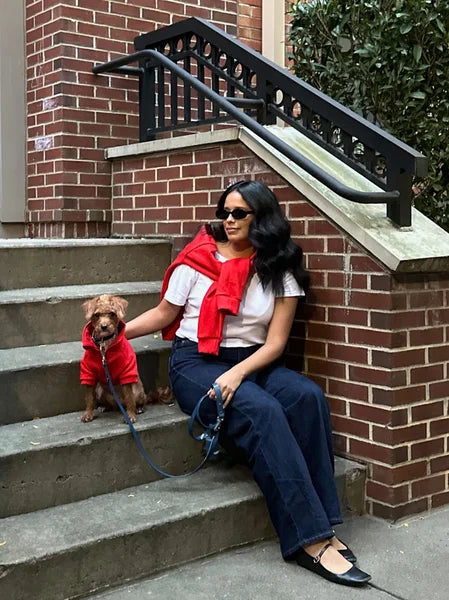 Woman sitting on steps with a small dog wearing a red coat, both sitting on a leash.