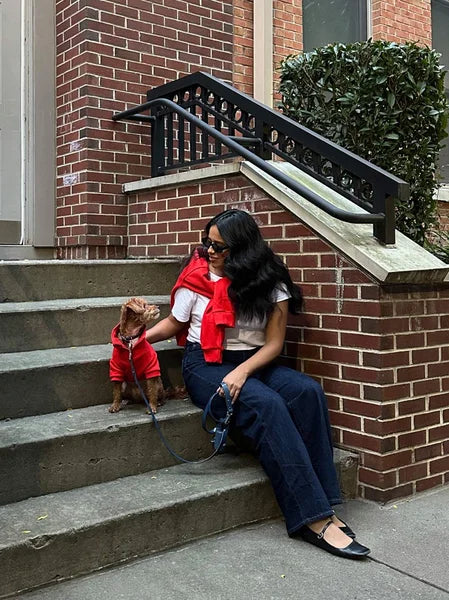 Woman sitting on steps with a small dog, both wearing red scarves, in front of a brick building.