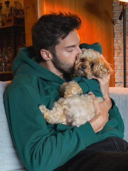 Man in a green hoodie holding a small dog indoors.