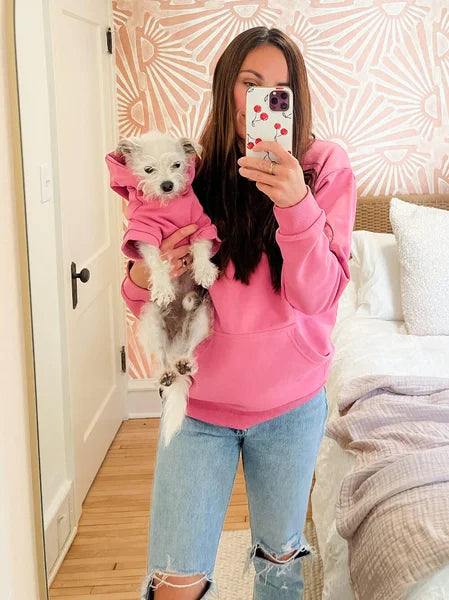 Person wearing a pink sweatshirt holding a small dog also in a pink outfit, standing in a room with a floral-patterned wall.