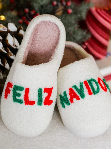 White slippers with 'Feliz Navidog' text in red and green on a blurred Christmas-themed background.
