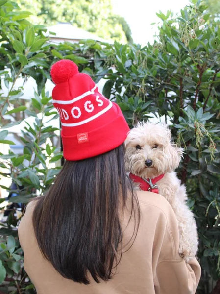 Person wearing a red 'DOGS' beanie with a dog on their back, surrounded by green foliage.