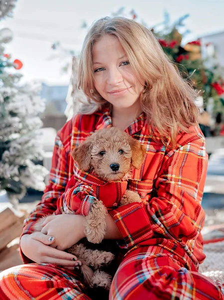 Person wearing red plaid pajamas holding a small dog in a snowy outdoor setting.