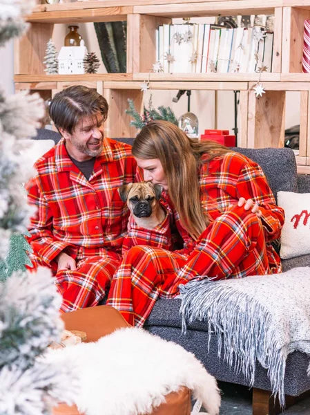Two people in red plaid pajamas sitting on a couch with a dog, surrounded by Christmas decorations.