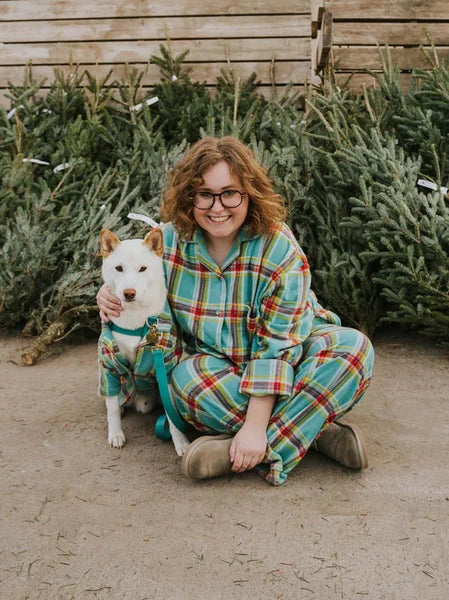 Woman in plaid pajamas sitting with a small dog in front of Christmas trees.