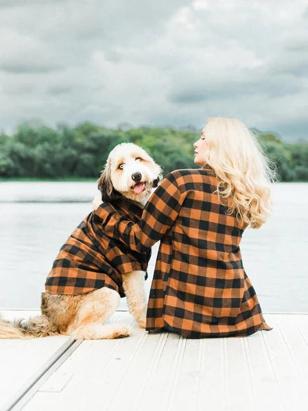 Woman in a checkered coat sitting on a dock with a dog, both looking out over water.