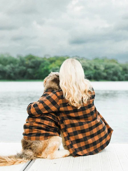 Person and dog sitting together on a dock by a lake, wearing plaid coats.
