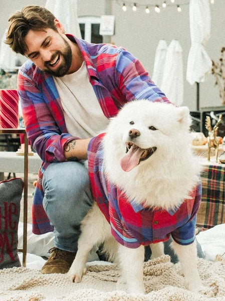 Man wearing a plaid shirt with a white dog also in a plaid outfit, both sitting on a carpeted floor.