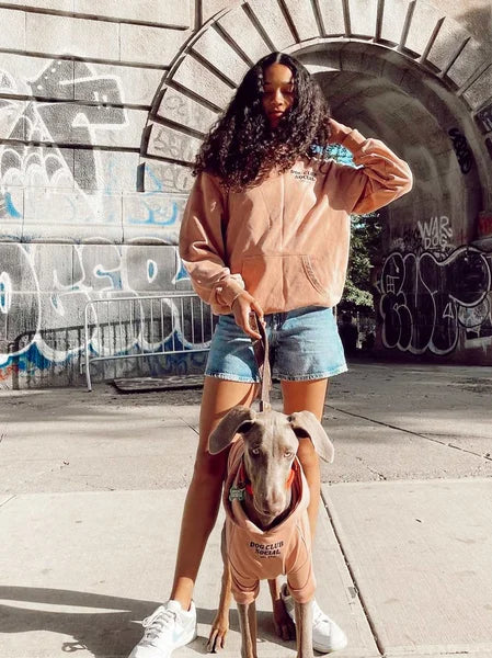 Person in a peach hoodie and denim shorts standing on a sidewalk with a dog, graffiti-covered wall in the background