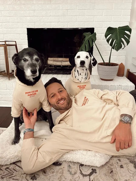 Man and two dogs wearing matching 'Dog Club' hoodies in a living room setting.