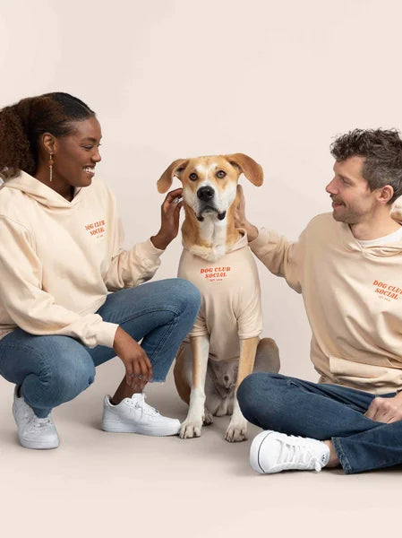 Two people in matching beige hoodies with a dog on a plain background