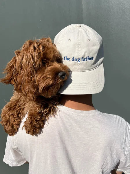 Person wearing a cap with 'the dog father' text and holding a brown dog against a gray background