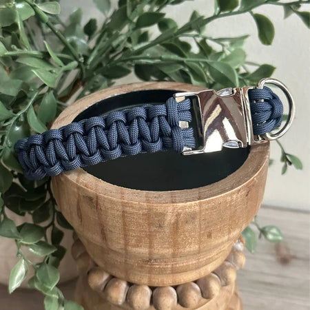 Blue paracord dog collar with metal clasp on a wooden stand with green leaves in the background
