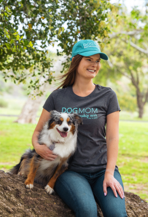 Woman wearing a 'Dogmom' shirt and cap sitting with a dog on a log in a park.