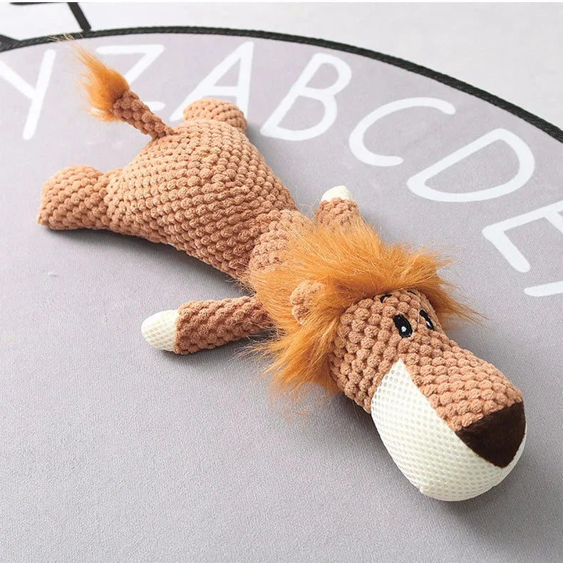 A Jungle Jamboree Chew Toy - Plush Squeaky Dog Toy Available in Wolf, Lion, or Elephant Designs shaped like a lion lies on its stomach on a gray mat. The lion has a brown knitted body, white paws, and a white snout. Its mane and tufted tail are made of orange fuzzy fabric. The mat underneath features white letters of the alphabet arranged in a circular pattern.