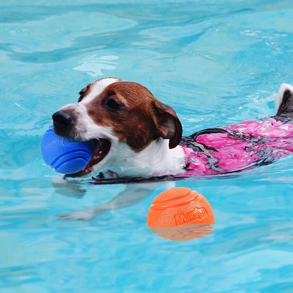 A dog with brown and white fur is swimming in a pool, holding a blue Durable Bouncy Dog Ball - Chew-Resistant Rubber for Outdoor Play & Training in its mouth. The dog is wearing a pink flotation vest. An orange ball floats nearby. Both balls appear textured, made of chew-resistant rubber by the brand 