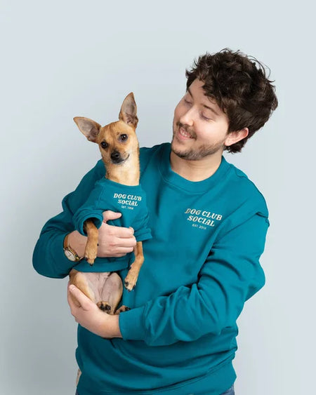 Man holding a small dog both wearing 'Dog Club Social' sweatshirts against a light gray background