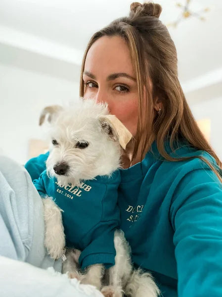 Woman holding a small white dog wearing a blue hoodie indoors.