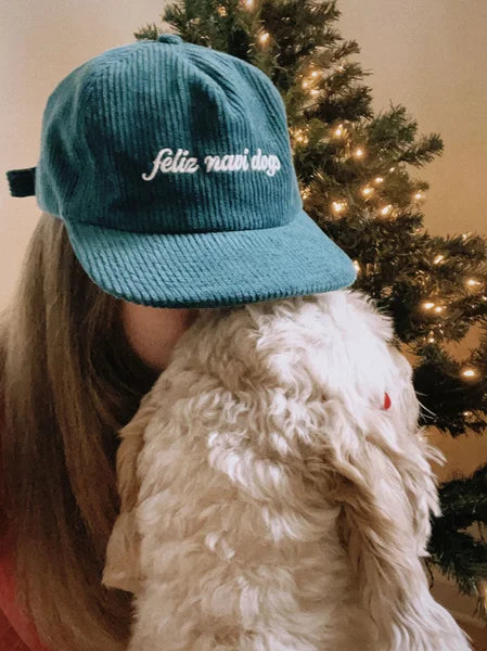 Person wearing a teal cap with 'felicidad' text, holding a fluffy dog in front of a decorated Christmas tree.