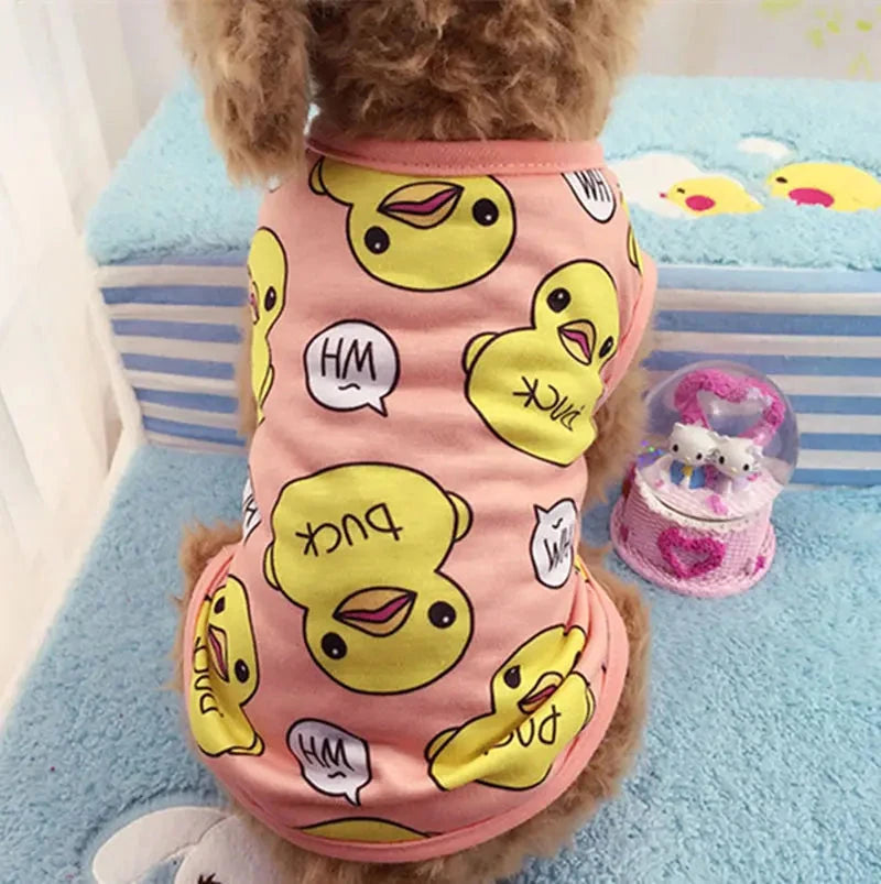 A small dog with curly fur sits on a blue towel, facing away from the camera. The dog is wearing Adorable Spring & Summer Puppy Pajamas - Soft, Stylish Dog Outfits for Small Breeds—a pink shirt adorned with yellow cartoon duck faces and speech bubbles. In the background, there's a small decorative snow globe with a pink base, adding to the charming scene.