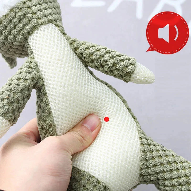 A hand is pressing the red dot on the belly of a green and white textured stuffed animal, crafted from non-toxic plush fabric. A red speaker icon in the top right corner indicates the toy has a sound feature activated by pressing the dot. The background is gray and slightly blurred.

Product Name: Jungle Jamboree Chew Toy - Plush Squeaky Dog Toy Available in Wolf, Lion, or Elephant Designs